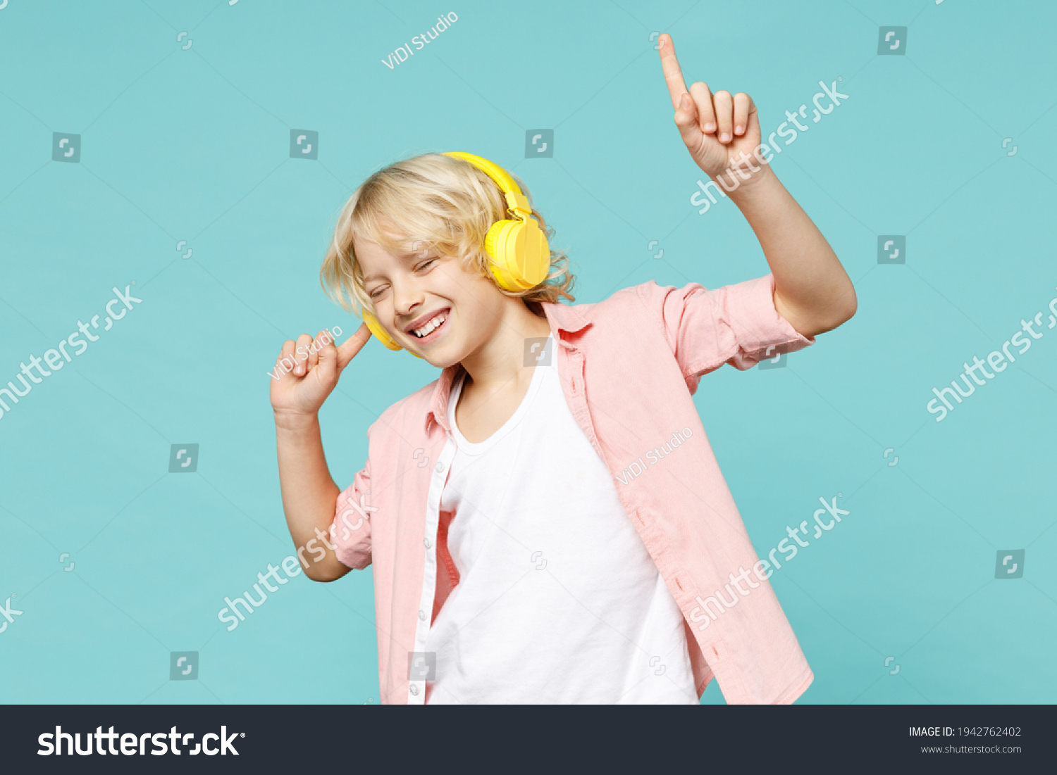 Funny little curly kid boy 10s years old in casual pink shirt listening music with headphones dancing isolated on blue turquoise color background children studio portrait. Childhood lifestyle concept