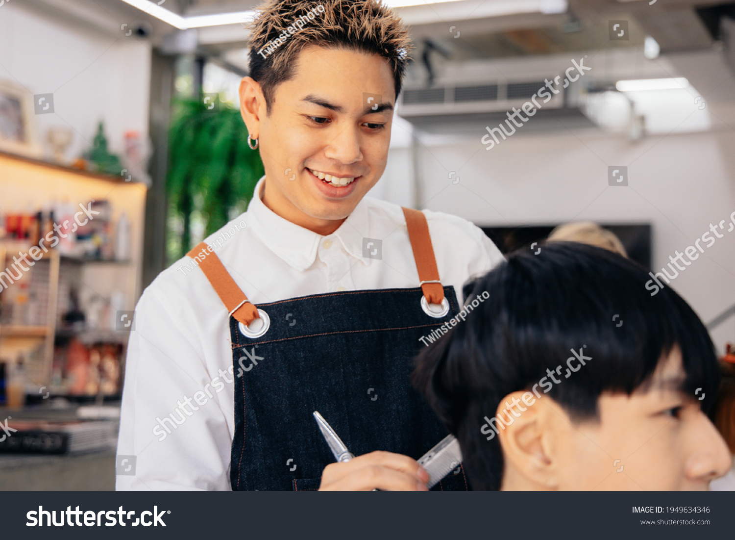 Happy young Asian male hairdresser smiling wearing apron in modern salon holding brush and scissors trimming hair of client