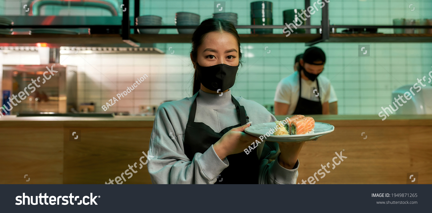 Portrait of young waitress wearing protective mask looking at camera and holding a plate with sushi rolls. Japanese cuisine in restaurant. Asian food concept. Web Banner