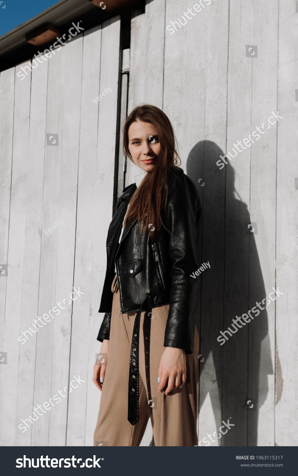 Smiling young woman in black leather jacket standing in front of white ...
