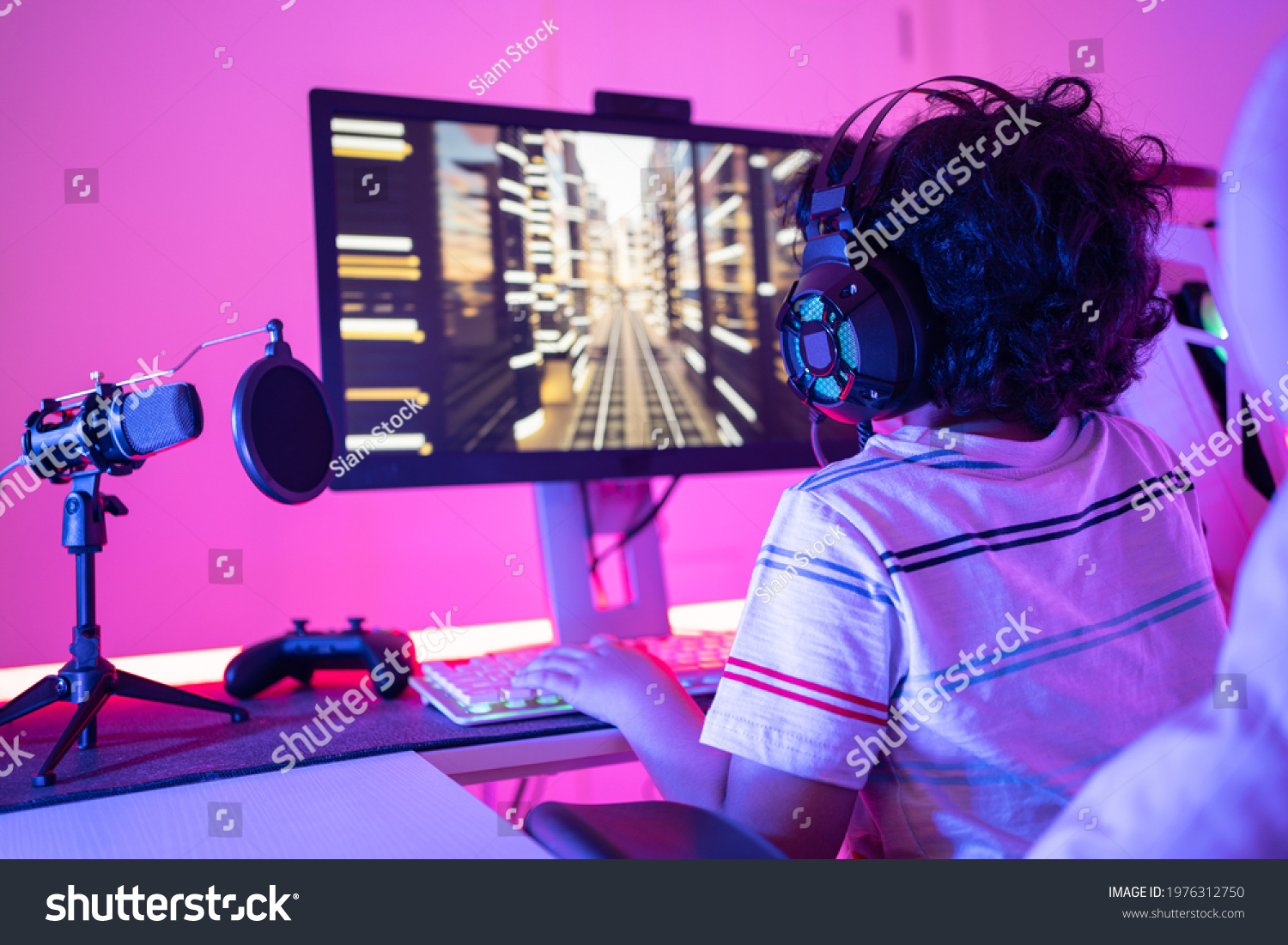 Back of cute curly-haired little Asian boy gamer wearing headphones sitting playing video games on a personal computer with RGB keyboard next to joystick and desktop microphone in a violet neon room.