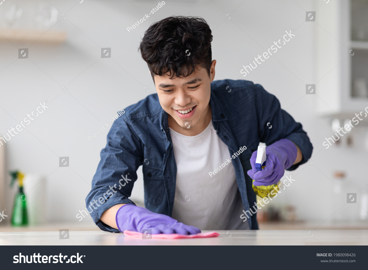 Cheerful asian guy cleaning kitchen table with spray and dust cloth  happy with his job  closeup  copy space. Smiling young chinese man house-keeping after moving to new apartment