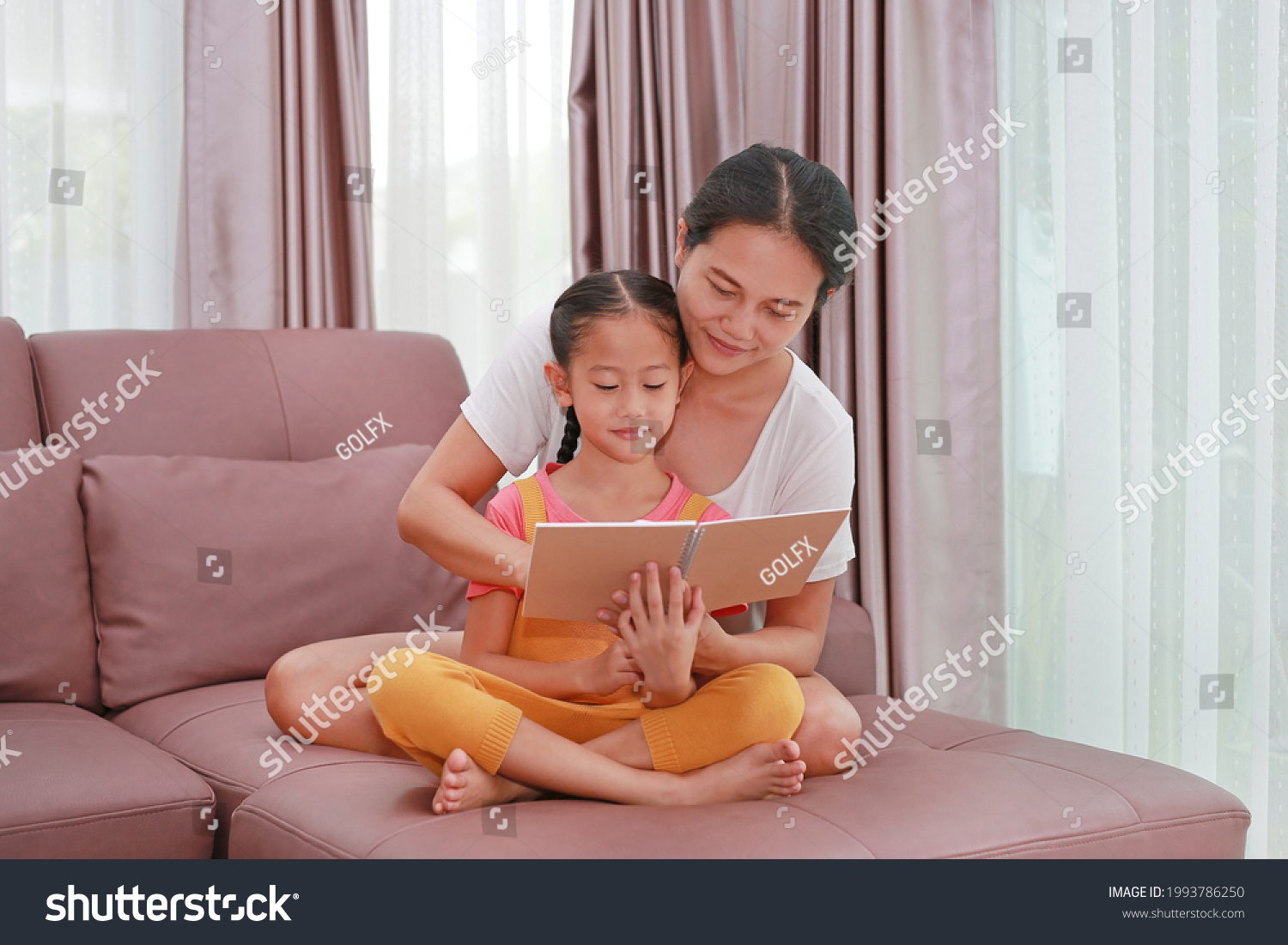 Asian mother and daughter reading a book on the sofa. Happy loving family at home