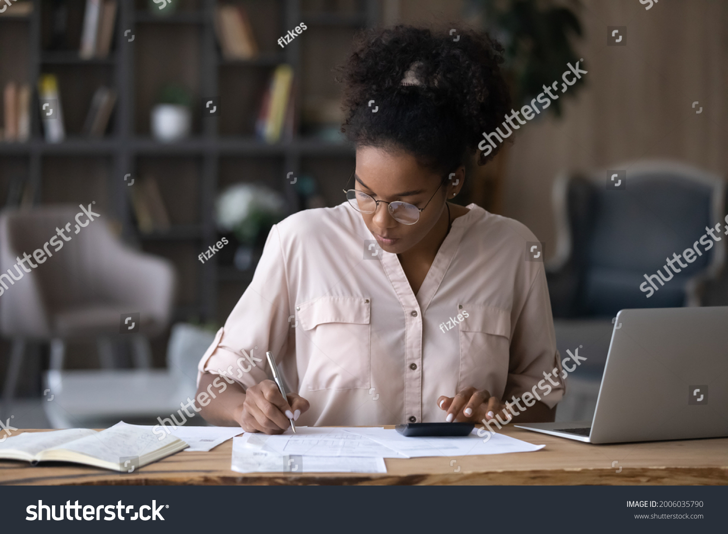Serious African American woman in glasses calculating expenses  using calculator  sitting at desk with laptop and financial documents  focused young female managing planning household budget