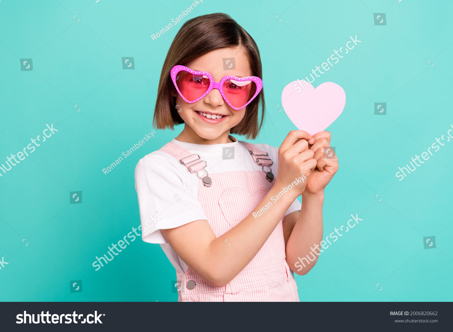 Photo of funny adorable school girl wear pink overall glasses smiling holding little heart isolated teal color background