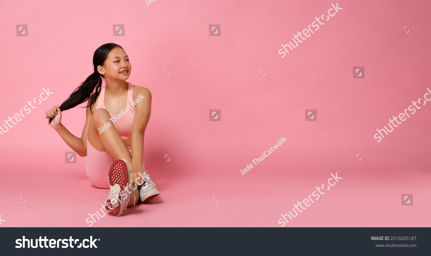 Sport Teenager girl sit and smile to left do fashion power poses. 12 years old Asian Youth Athlete kid wear Pastel Pink Fitness cloth pants over pink background full length