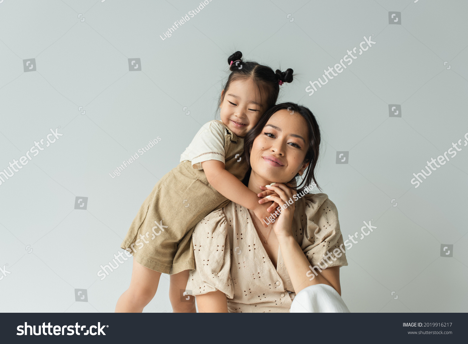 cheerful asian toddler daughter hugging happy mother isolated on gray