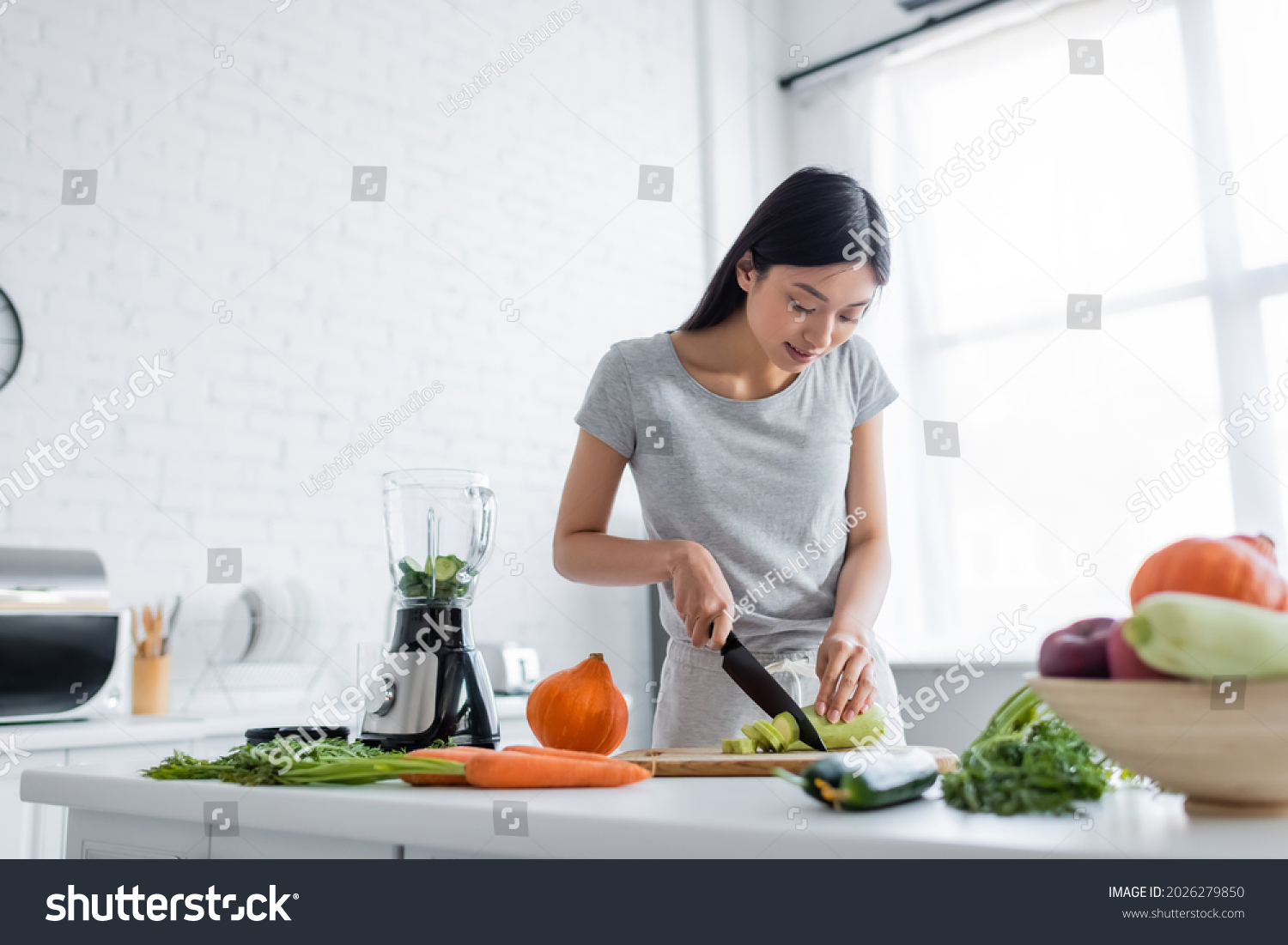 young asian woman cutting zucchini near raw vegetables and electric shaker in kitchen