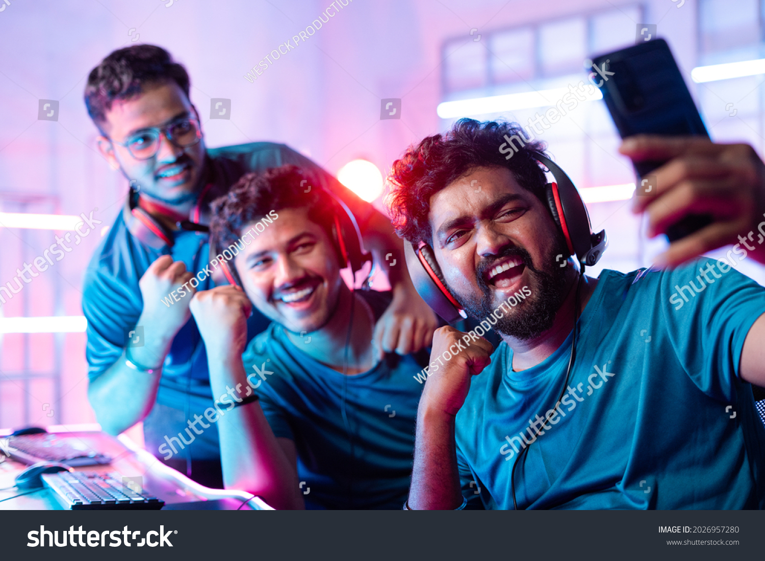 Team of happy cheerful professional gamers with headphones vlogging  live streaming  or taking selfie on mobile phone in front of monitors during esports video gaming tournament