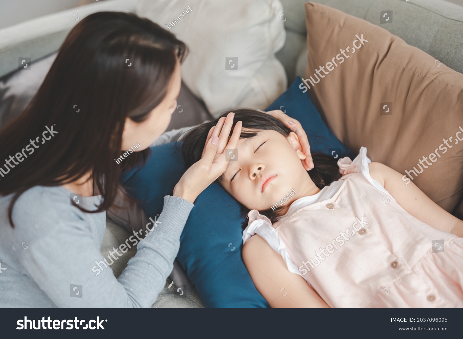 Sick Asian girl daughter sleeping on the couch with fever while mother checking temperature on her forehead