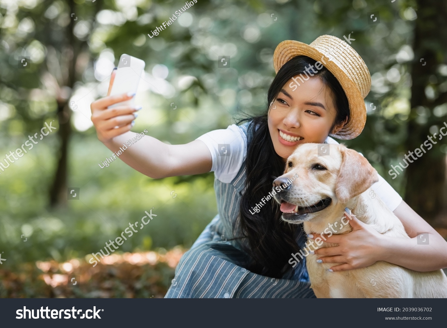 joyful asian woman in straw hat taking selfie with yellow labrador in park