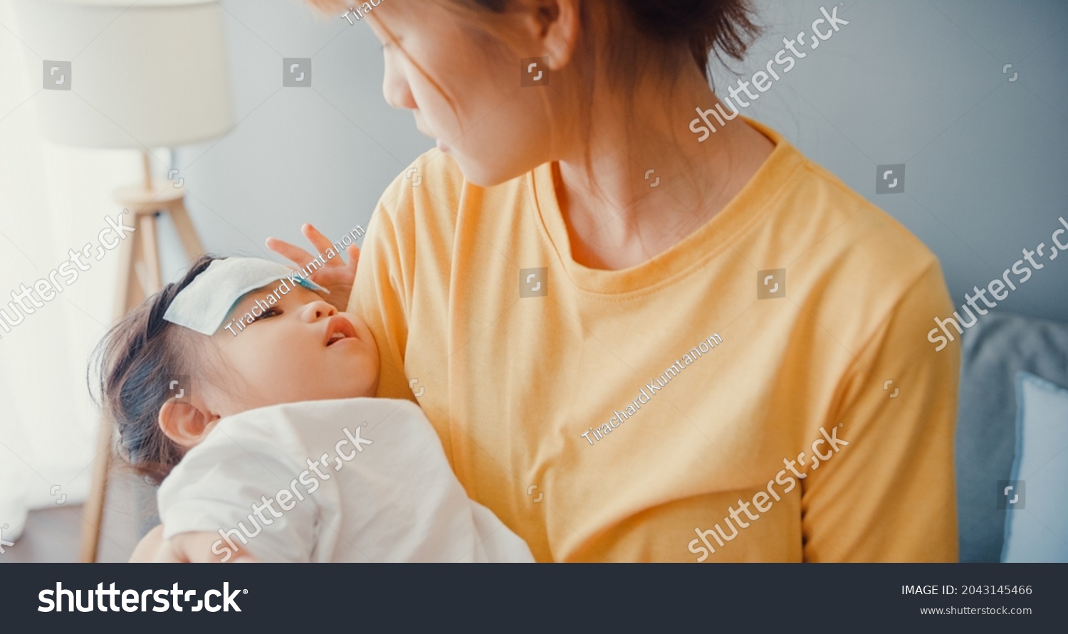 Happy cheerful Asia family mom holding sick toddler girl in arms and attach the antipyretic gel on baby forehead walk in living room at house. Medical care insurance treatment and healthcare concept.