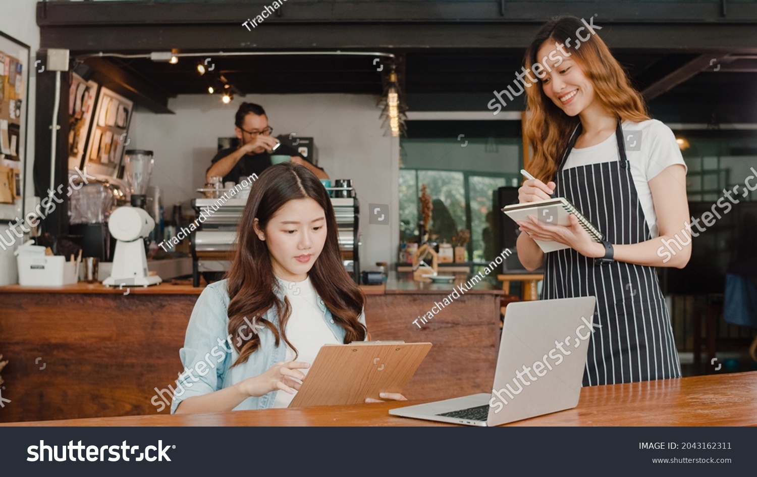 Cheerful Asian lady waitress with notebook taking receive order menu from young client girl at urban cafe. Young Asian freelance women working on laptop at coffee shop. Owner small business concept.