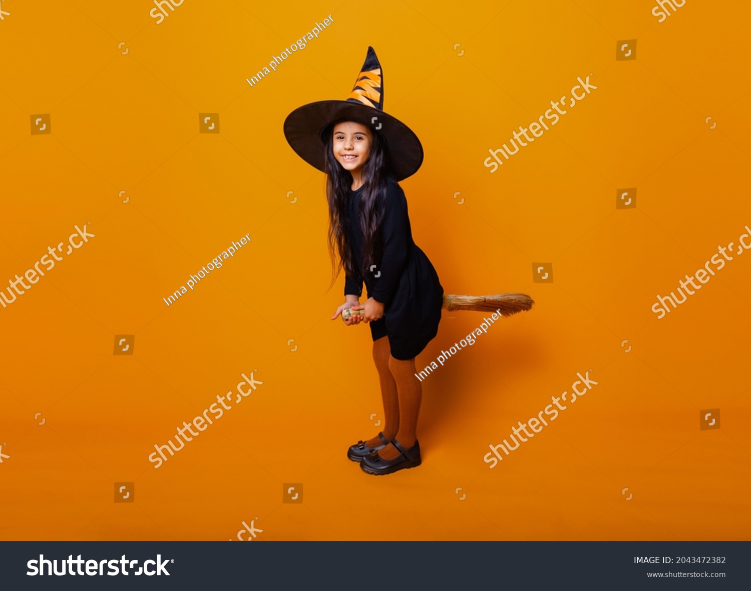 Little girl dressed as a Halloween witch in a black dress and hat flies on a broomstick on a yellow background.