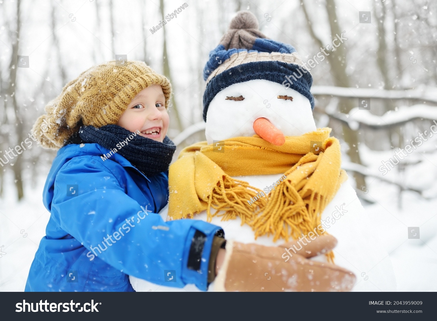 Little boy building snowman in snowy park. Child embracing snowman wearing hat and scarf. Active outdoors leisure with family with children in winter. Kid during stroll in a snowy winter park