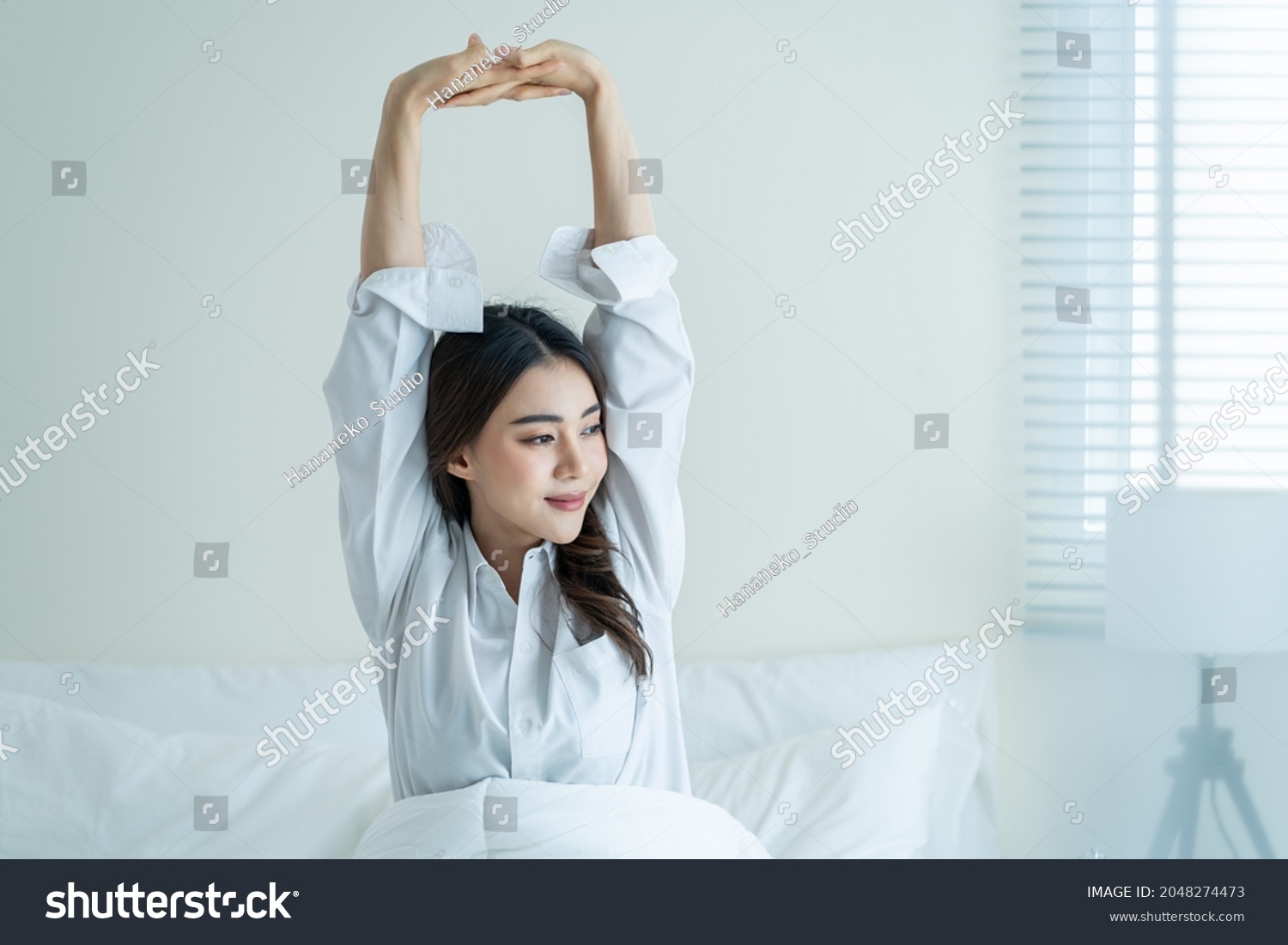 Asian beautiful girl in pajamas wake up in the morning with happiness. Attractive young woman smiling feel happy and relax then stretching body after getting up from sleep on bed in bedroom at home.
