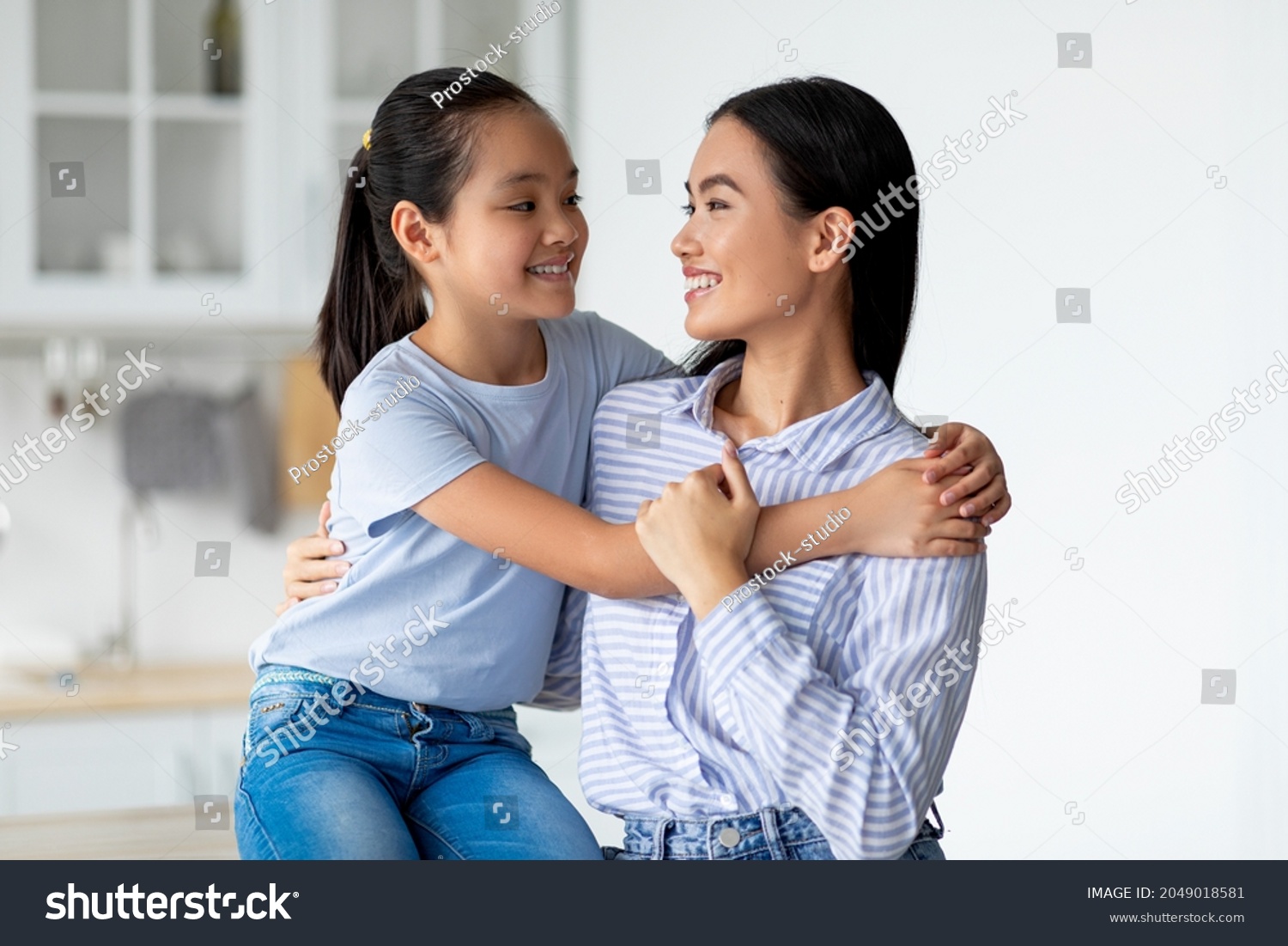 Mother daughter love. Happy asian lady hugging with her child  sitting in kitchen interior. Smiling girl embracing her mom at home  greeting with mother's day  copy space