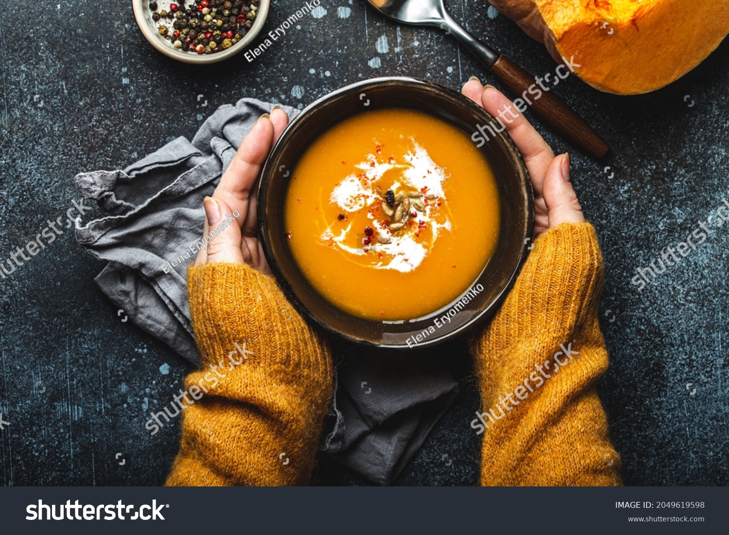 Female hands in yellow knitted sweater holding a bowl with pumpkin cream soup on dark stone background with spoon decorated with cut fresh pumpkin  top view. Autumn cozy dinner concept 
