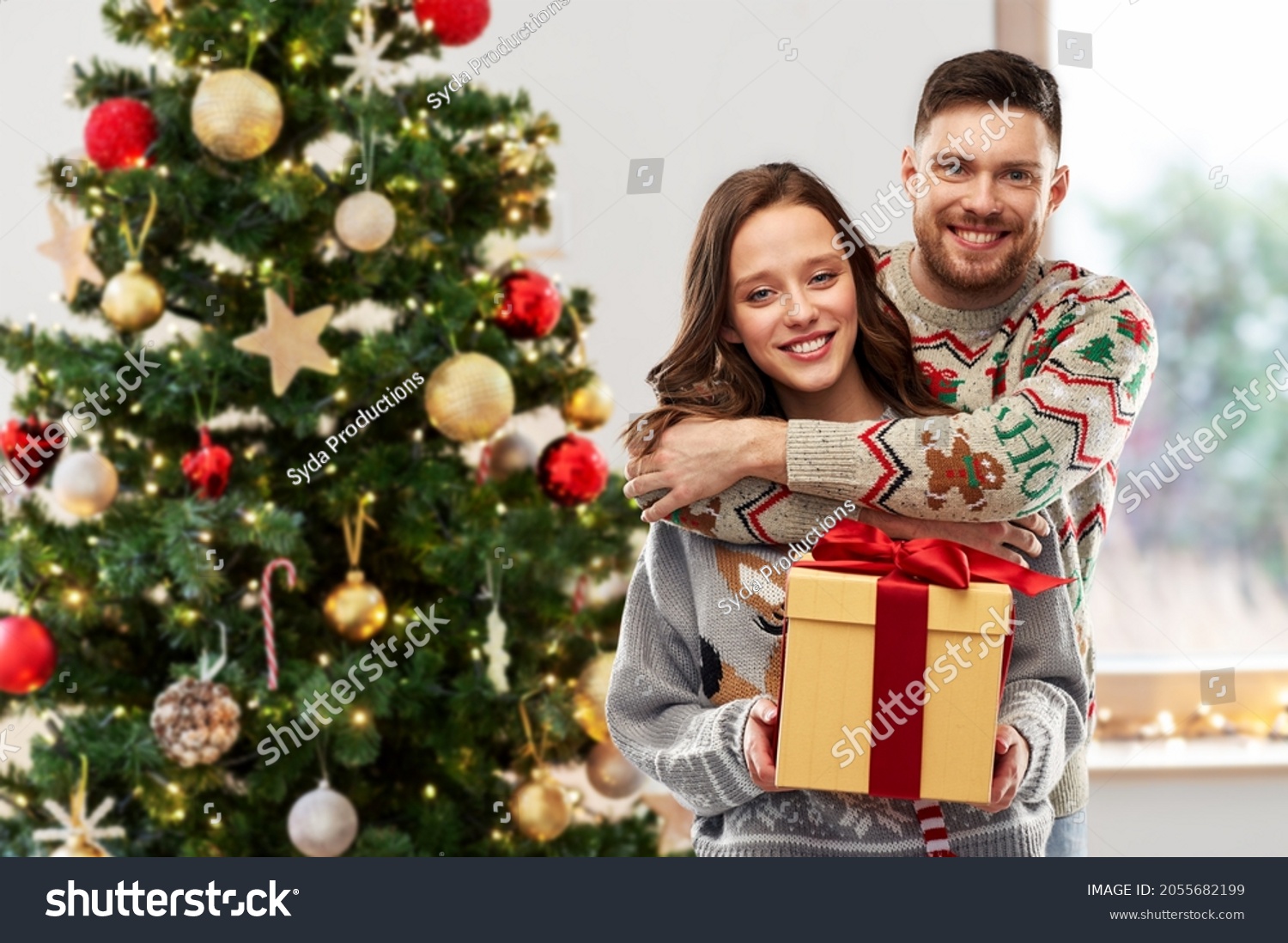 people and holidays concept - portrait of happy couple with gift at ugly sweater party over christmas tree on background