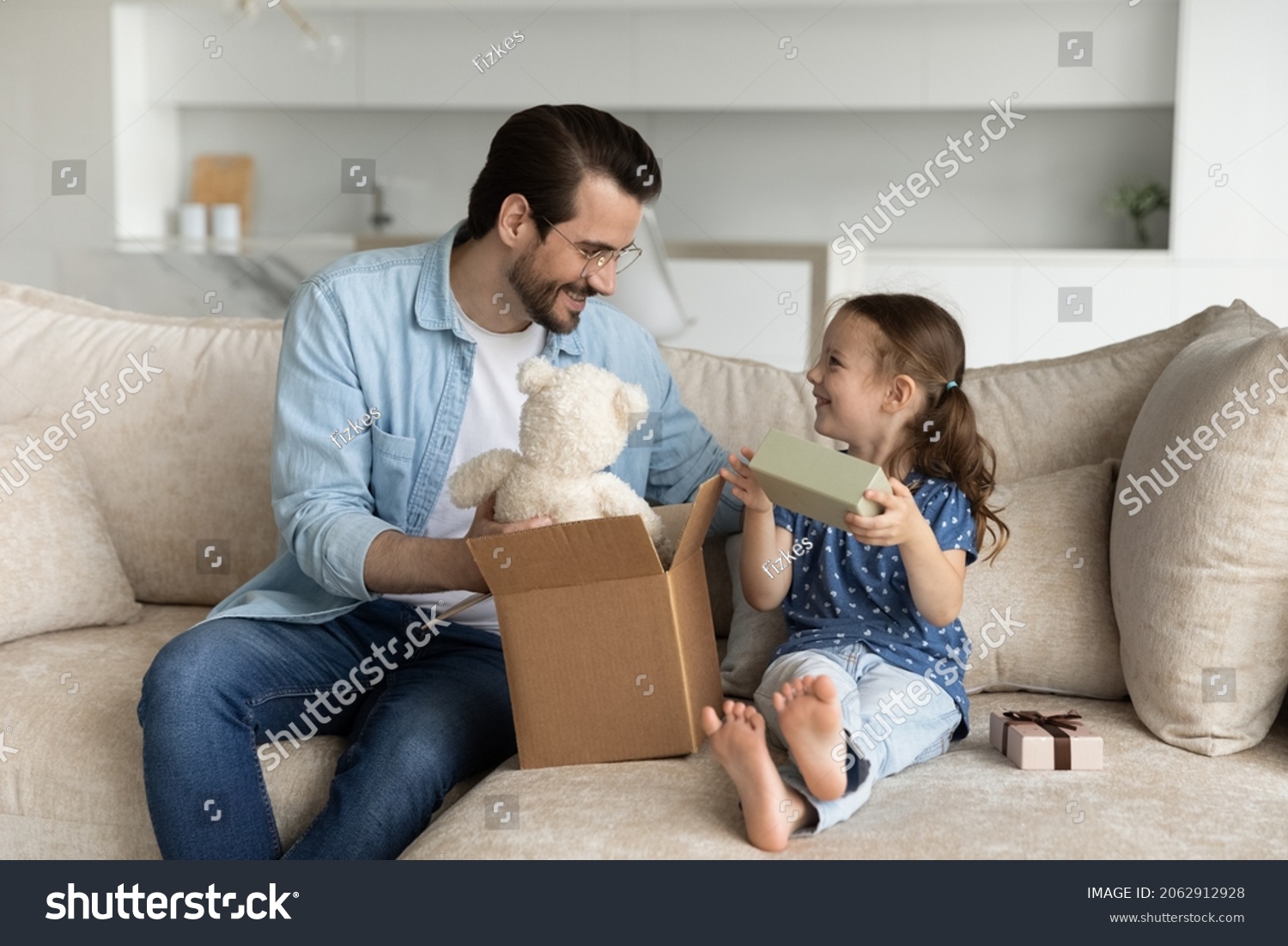 Happy young father unboxing parcel with gifts for small cute kid daughter sitting together on cozy sofa in living room satisfied with fast delivery purchase shipment or high quality fluffy toy.