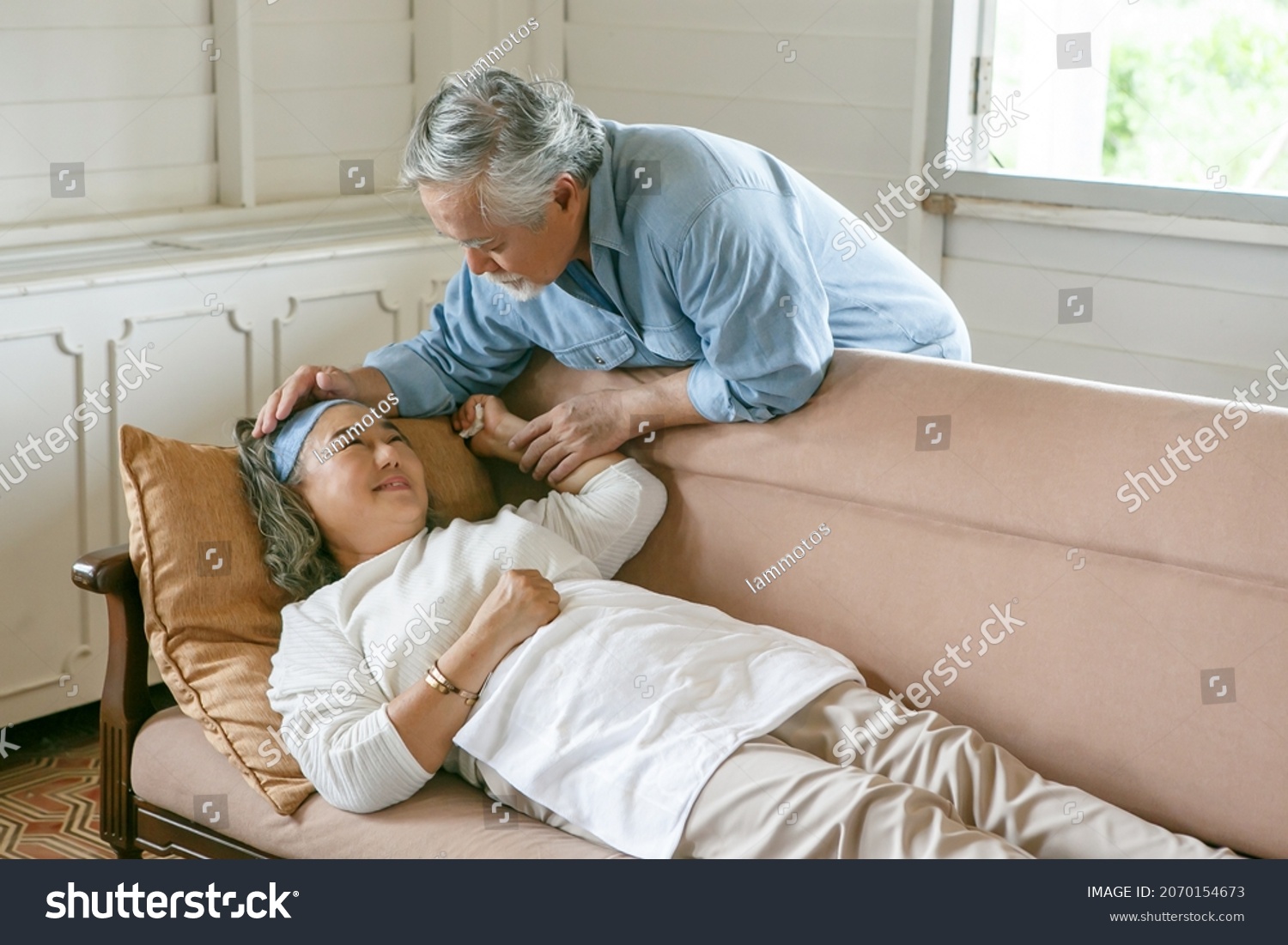 happy old asian husband take care and embracing elderly wife suffering pain of sick lying down on sofa in living room at home . Lovely asia senior couple in love support together on couch