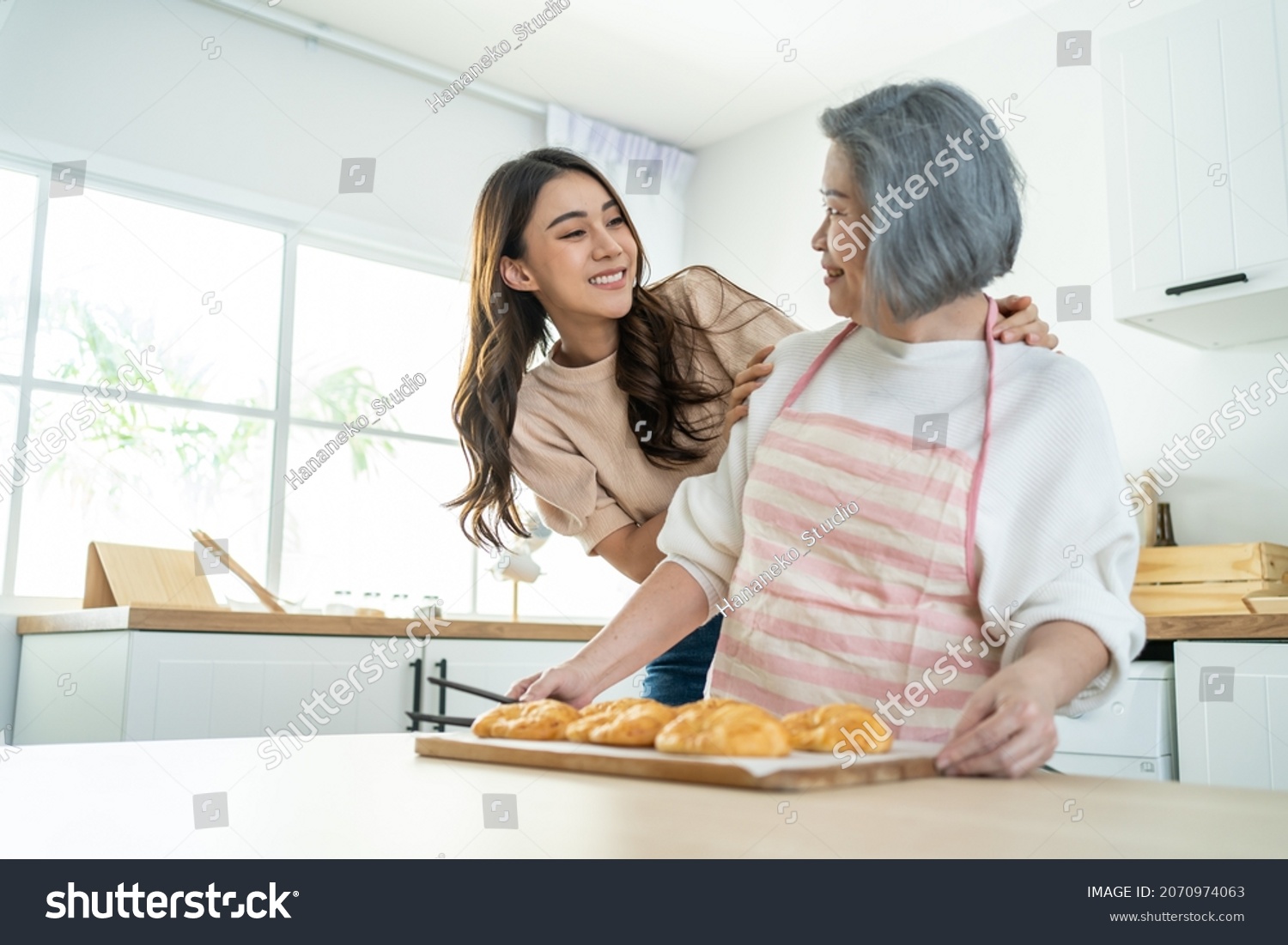 Asian lovely family young daughter look to old mother cook in kitchen. Beautiful female enjoy spend leisure time and hugging senior elderly mom bake croissant on table in house. Activity relationship