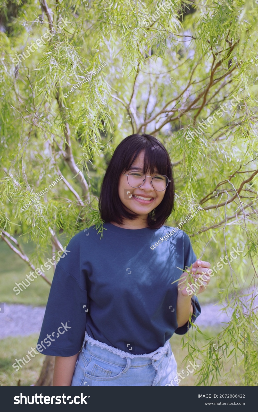 asian teenager toothy smiling and standing under green leaves tree