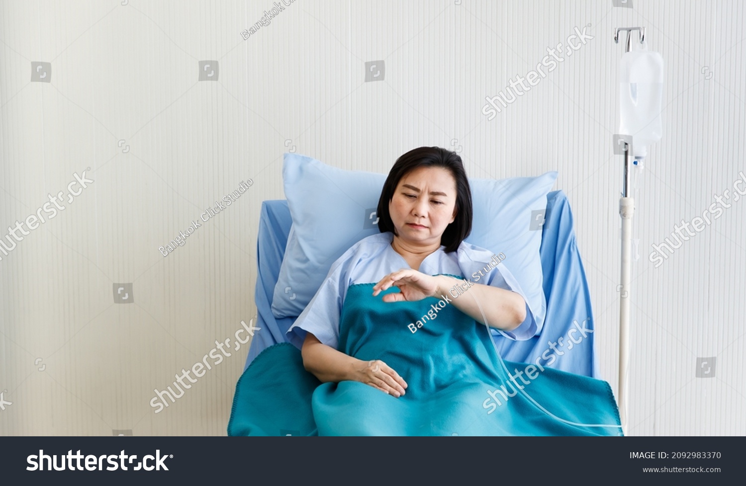 Asian senior stressed depressed upset doubtful sick female patient in blue hospital uniform laying down on bed alone looking at hand with injecting needle receiving normal saline solution from bag.