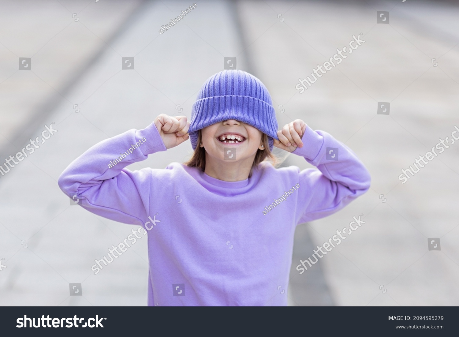 Cute little caucasian girl eight years old with blonde hair smiling outdoor. Kid wearing stylish shirt and knitted hat violet color. Trendy color of the 2022 year very peri