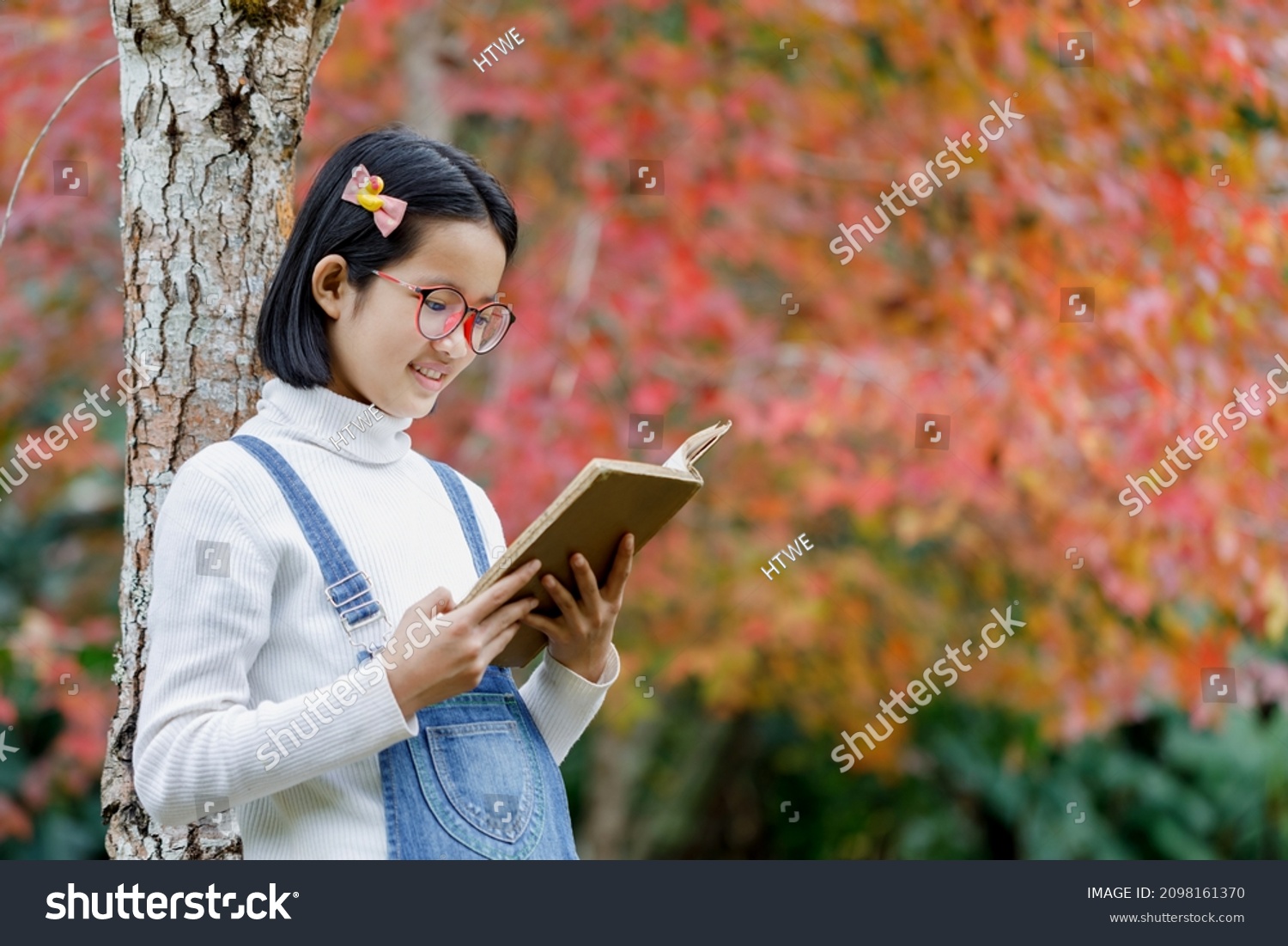 Smiling asian cute little girl with glasses reading book under maple tree