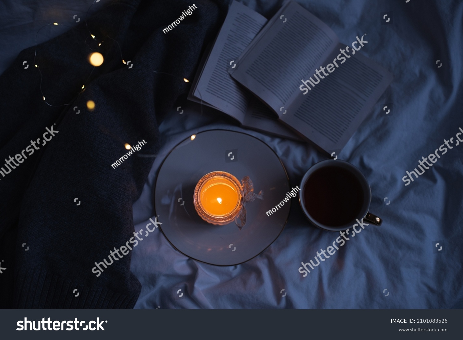 Burning scented candle on ceramic tray with open paper book and knit sweater cloth in bed. Winter cozy hygge atmosphere with cup of black coffee at home. Coziness. 