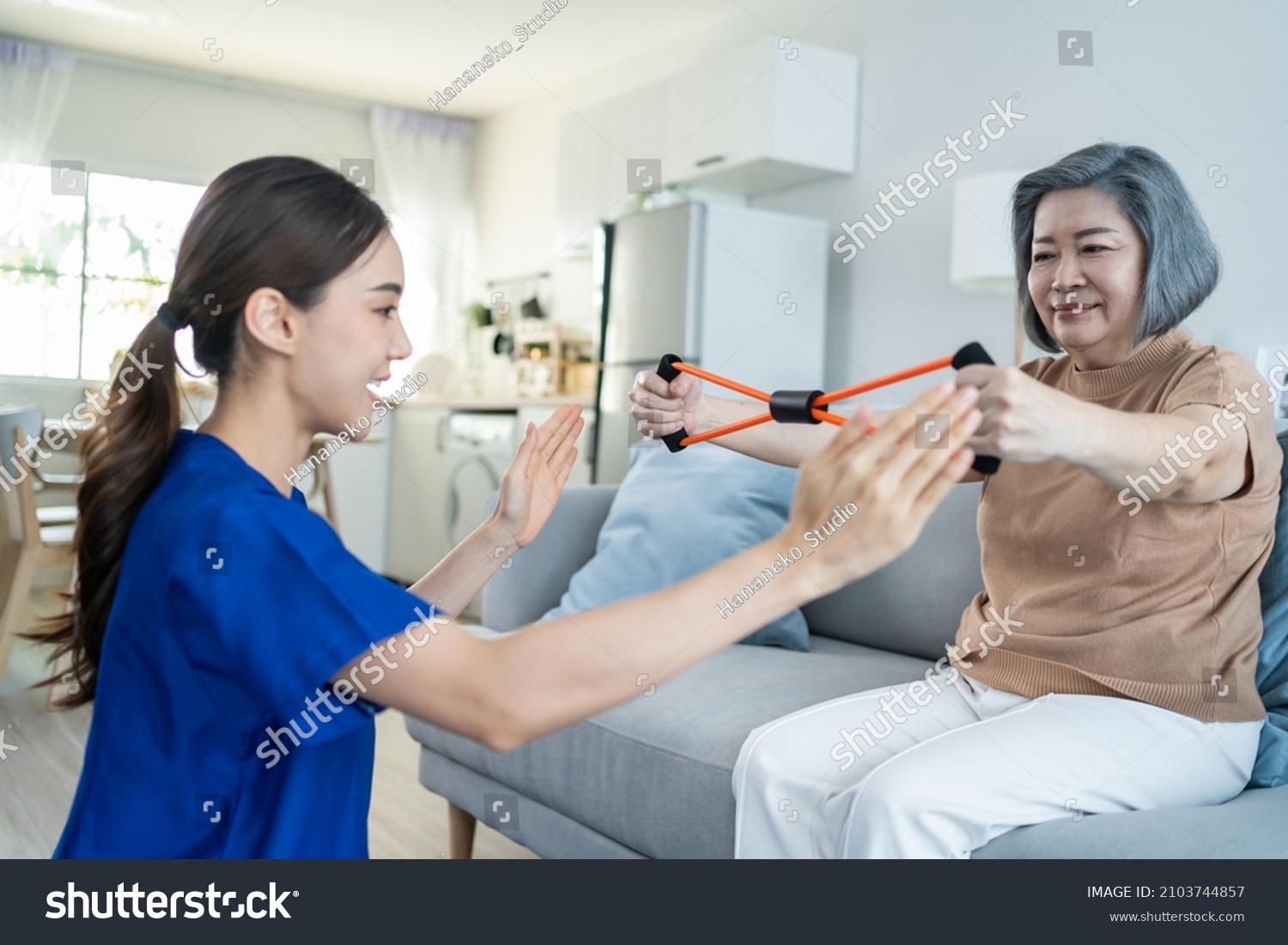 Asian mature older woman doing physiotherapist with support from nurse. Senior elderly female patient sitting on sofa in living room use resistance stretch band exercise with caregiver in nursing care