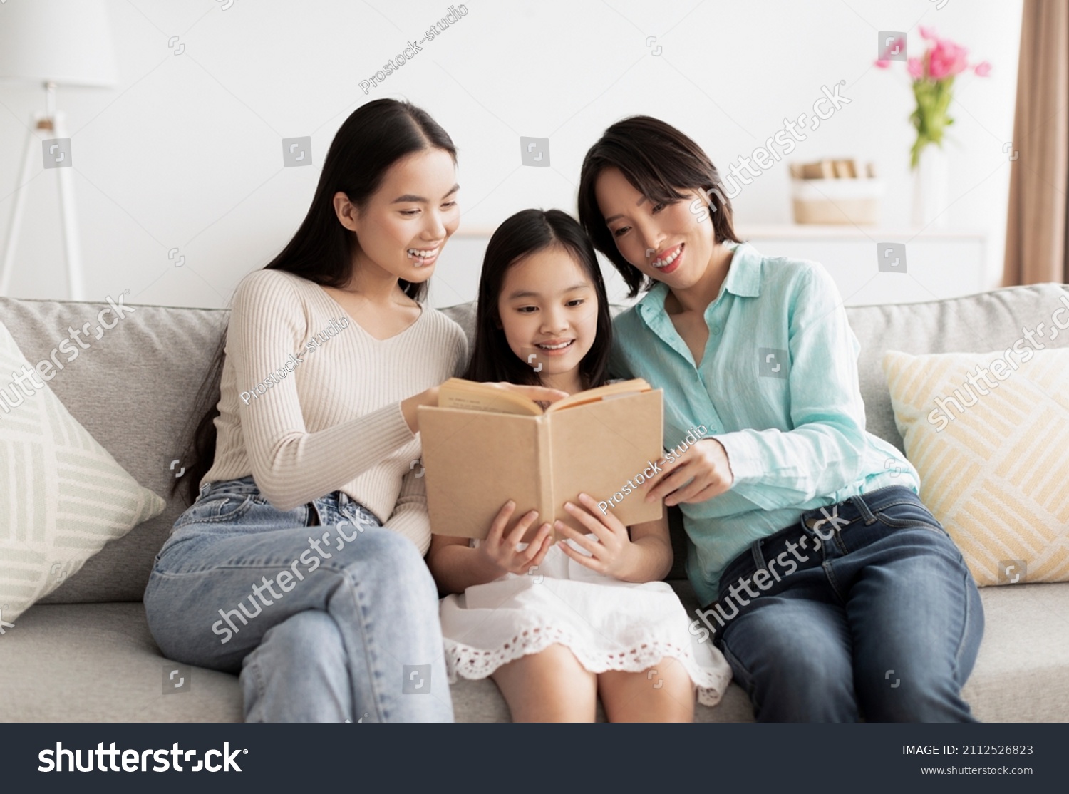 Cute little Asian girl reading book with her mommy and granny on couch at home. Affectionate multi generation family enjoying interesting story together in living room