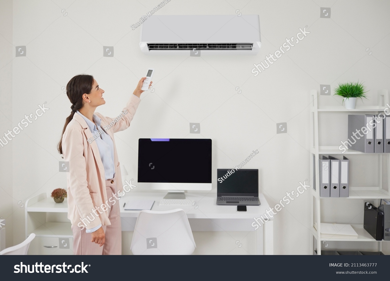 Happy office worker or business lady turns on AC. Smiling young woman in suit standing in office interior and switching wall air conditioner with remote to enjoy cool breeze on hot summer day at work