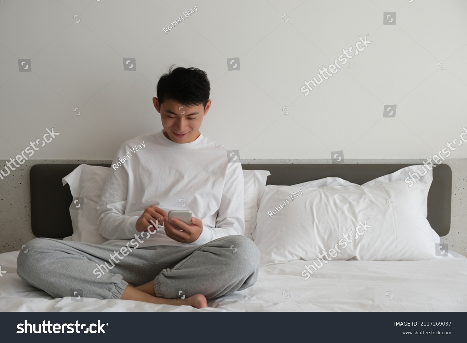 Smiling Asian young man sitting on bed  using and looking at phone at home