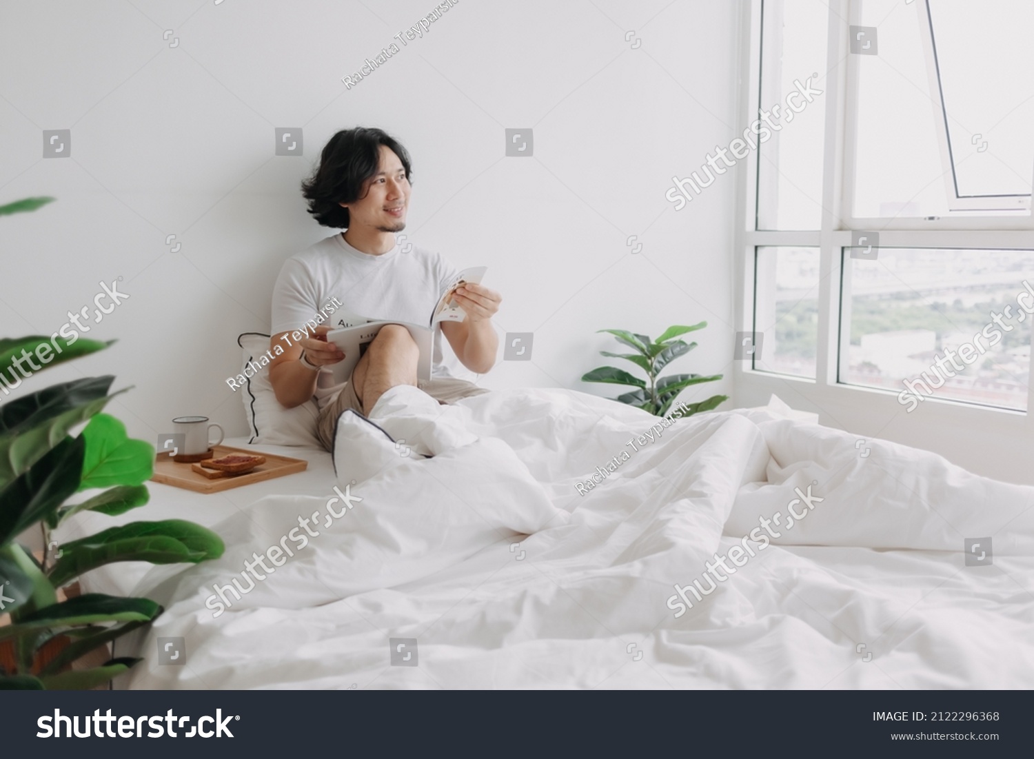 Asian man reading book on the bed with breads and coffee in his apartment.