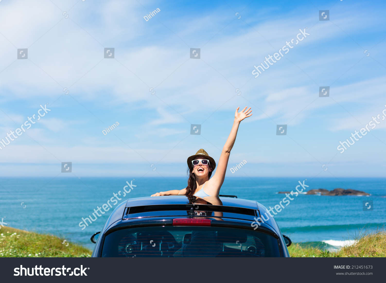 Joyful woman waving on summer car travel vacation to the coast. Brunette girl having fun leaning out vehicle sunroof towards the sea.