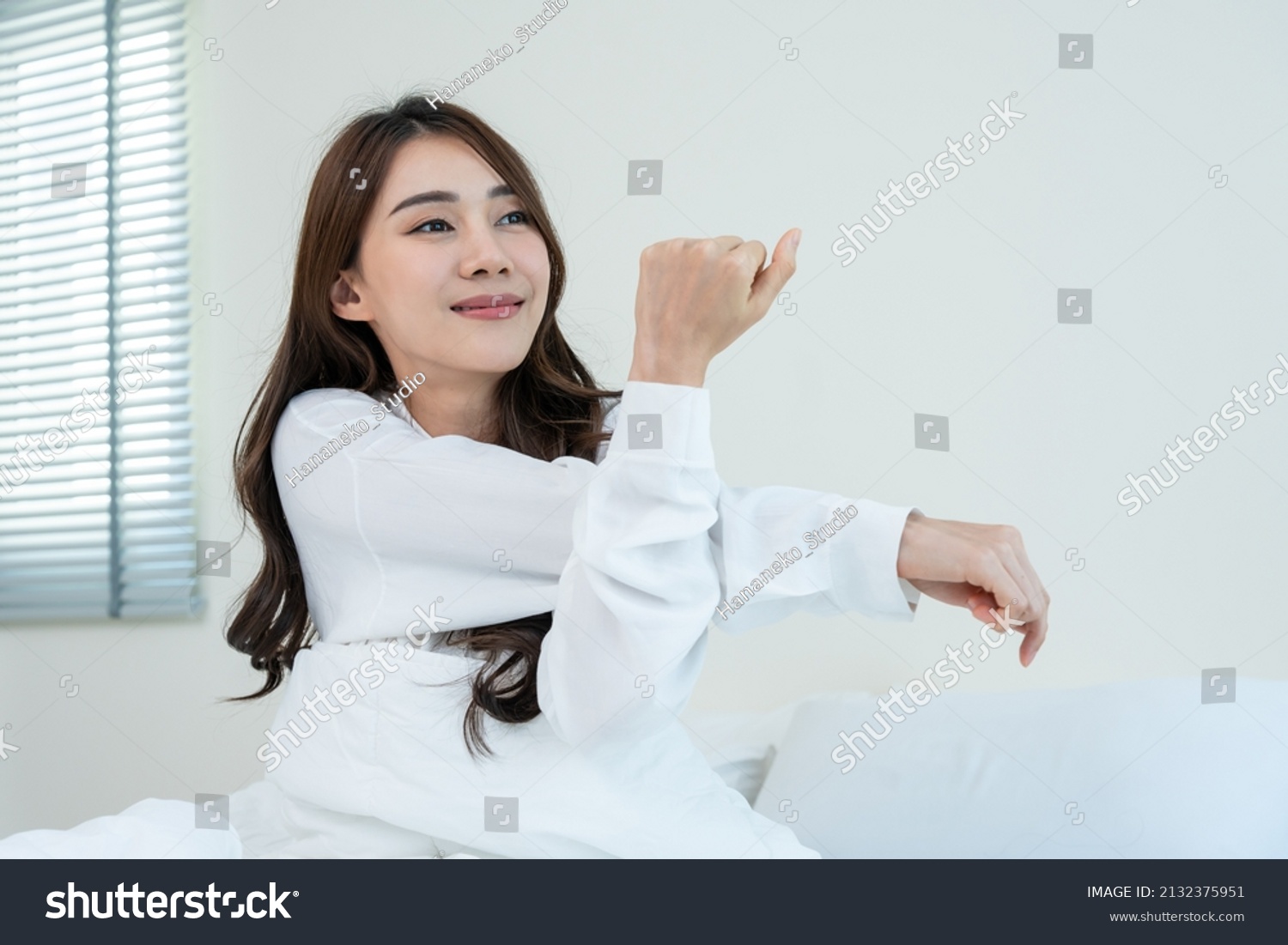 Asian beautiful girl in pajamas wake up in the morning with happiness. Attractive young woman smiling feel happy and relax then stretching body after getting up from sleep on bed in bedroom at home.