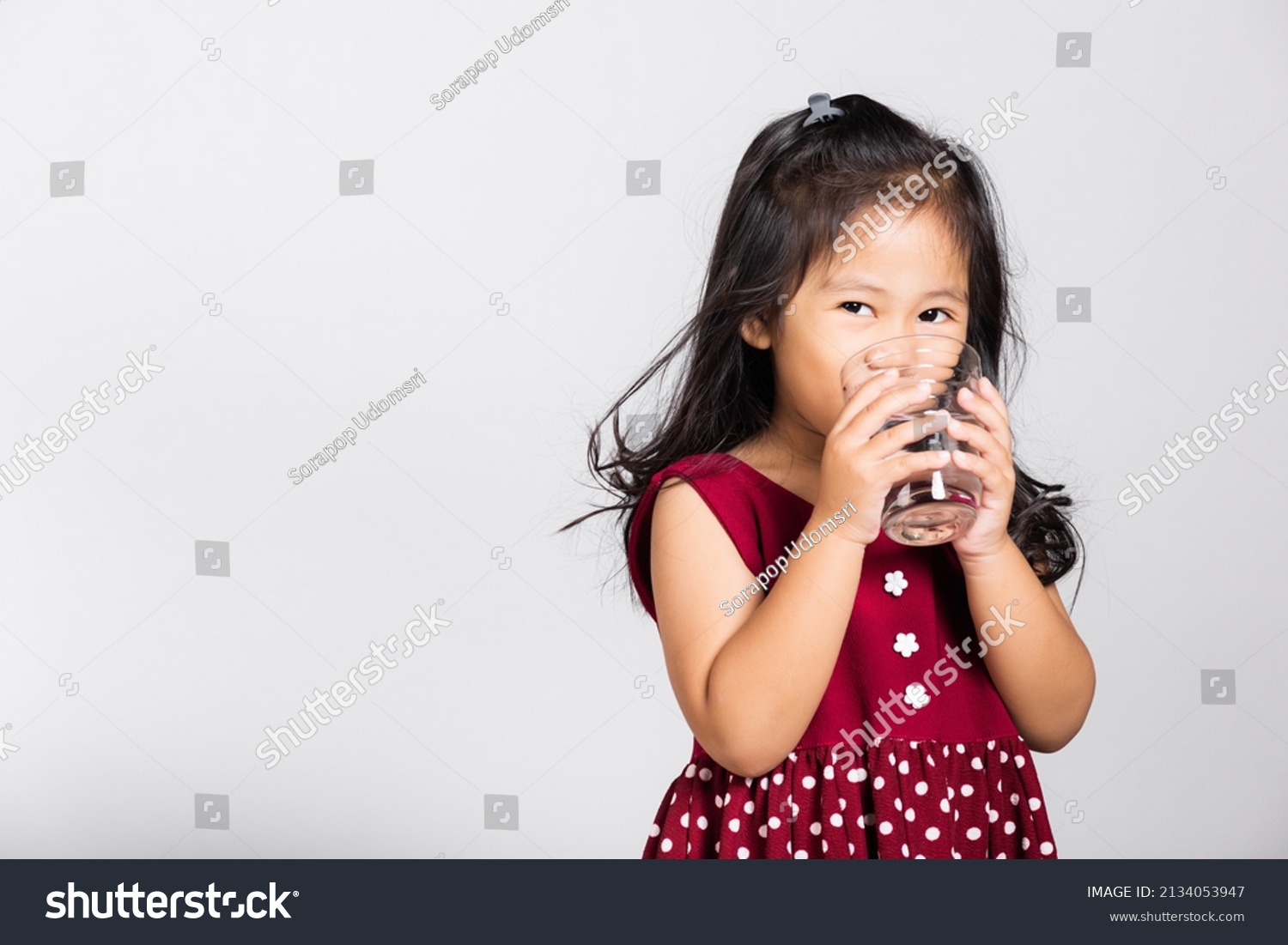 Little cute kid girl 3-4 years old smile drinking fresh water from glass in studio shot isolated on white background Asian children preschool Daily life health