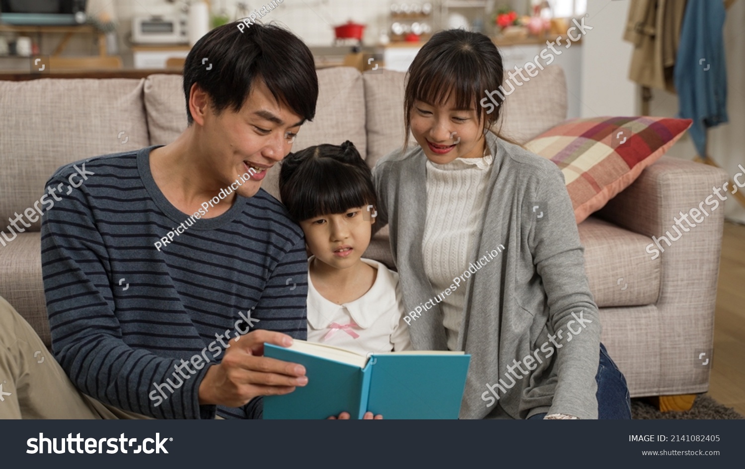 cheerful asian family of three enjoying reading together in the living room at home. the smiling father is having fun telling story with a book in hand.