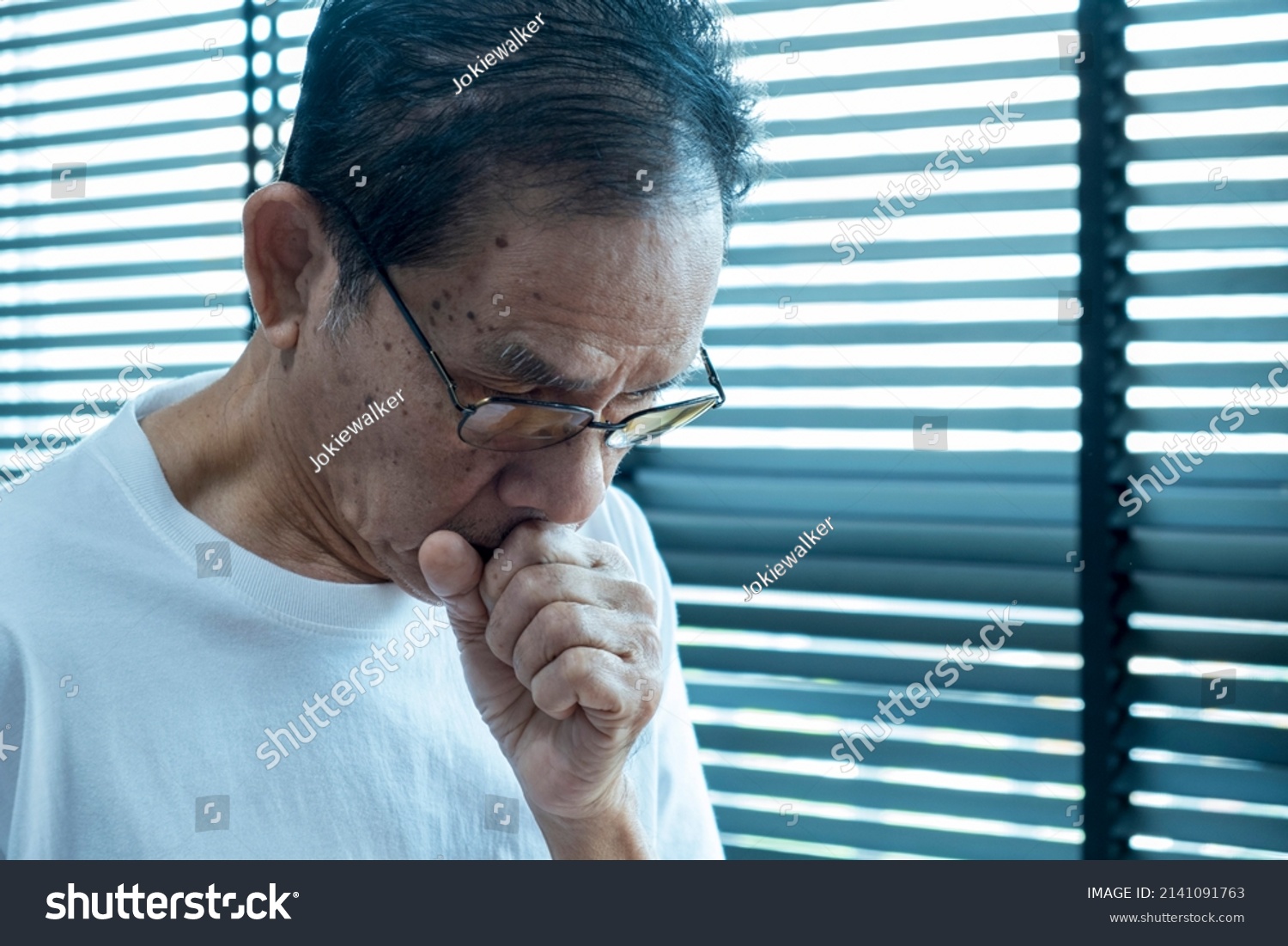 senior asian man coughing wearing glasses after waking up in the morning in the bedroom wearing a white shirt covering his mouth with hands