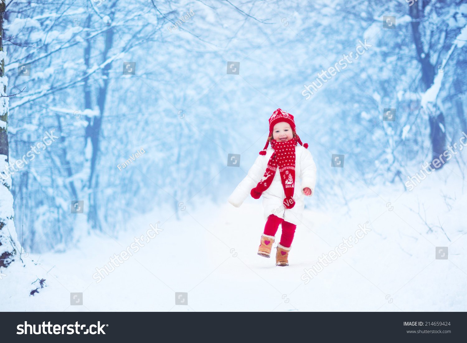 Happy laughing toddler girl wearing a white down jacket and red knitted hat and scarf playing and running in a beautiful snowy winter park on Christmas day