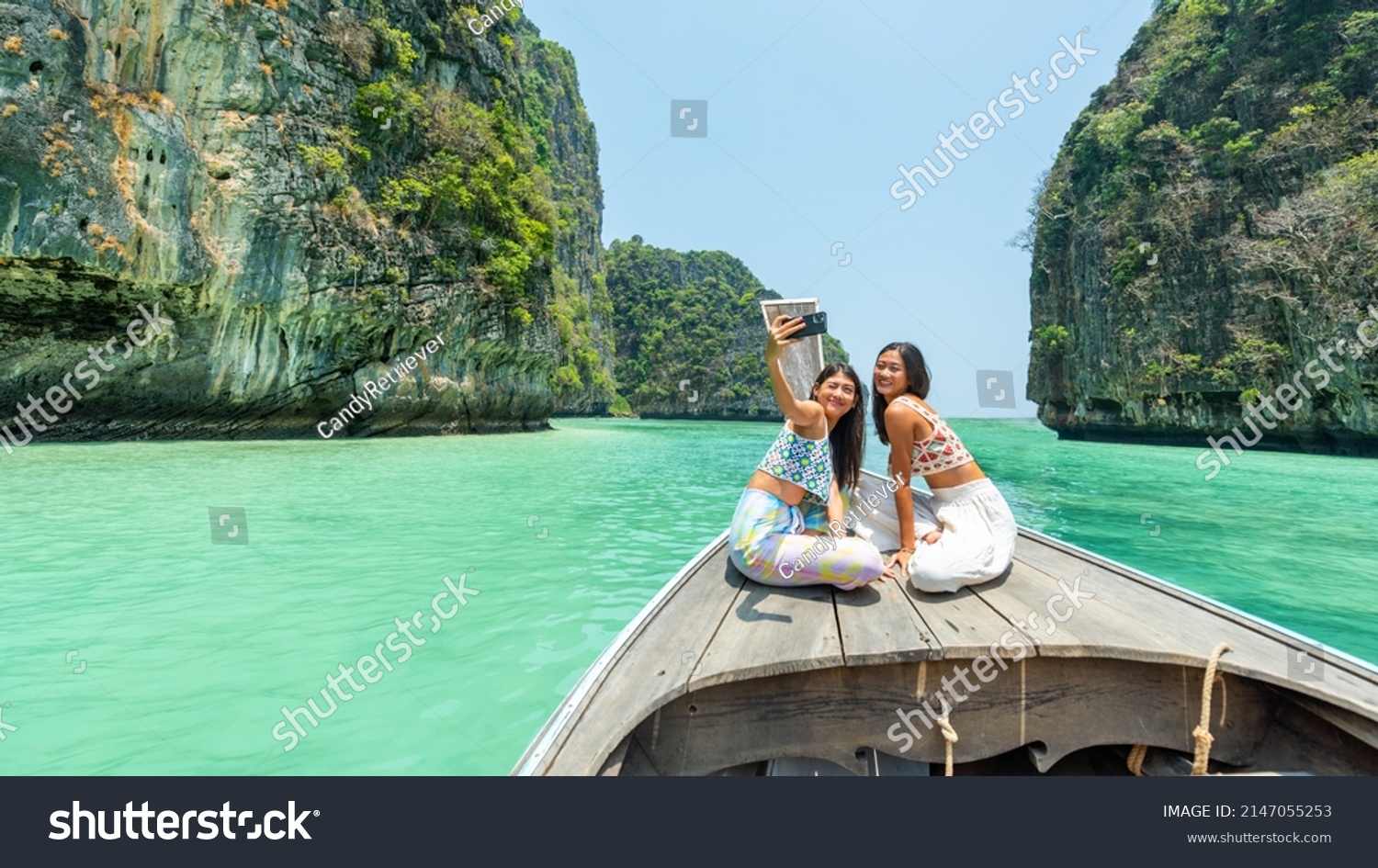 Young Asian woman friends using mobile phone taking selfie together while travel on boat passing island beach lagoon in sunny day. Happy female enjoy and fun outdoor lifestyle on summer vacation trip
