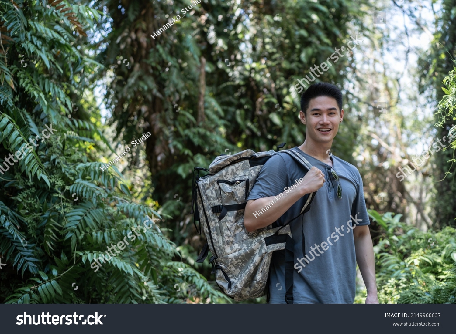 Portrait of Asian handsome man backpacker traveling alone in forest. Attractive male traveler walking in nature wood with happiness and fun during holiday vacation trip then smiling looking at camera