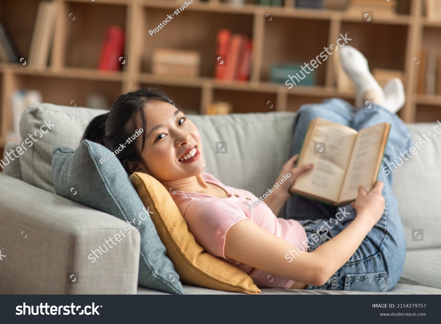 Cheerful asian lady resting on couch in living room reading book while spending time at home on weekend looking at camera and smiling. Hobby domestic entertainment