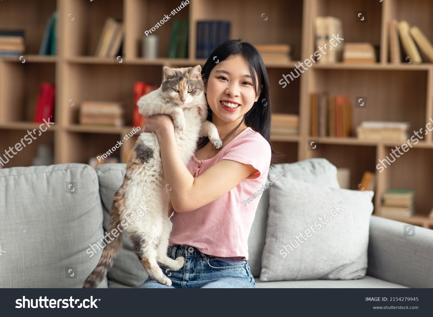 Pretty korean lady posing with her beautiful cat sitting on sofa in living room at home. Happy chinese woman spending time with lovely pet holding kitten and smiling at camera copy space