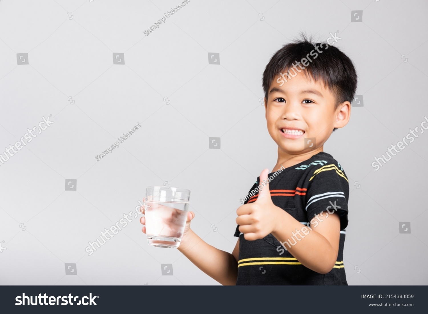 Little cute kid boy 5-6 years old smile drinking fresh water from glass and show thumb up finger for good sign in studio shot isolated on white background  Asian children preschool  Daily life health