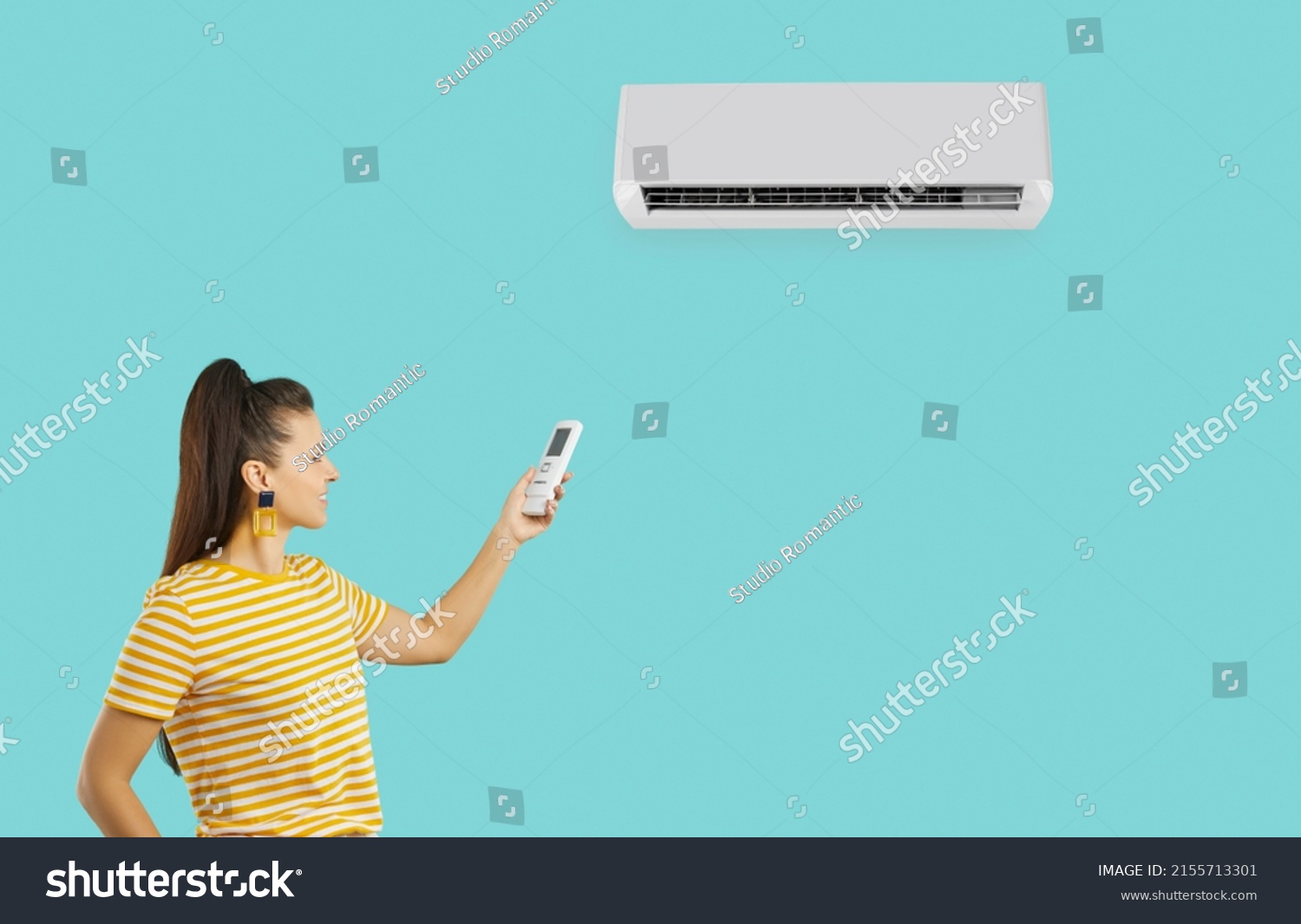 Happy woman for comfort during summer heat uses modern air conditioner isolated on turquoise background. Smiling millennial woman in yellow T-shirt uses remote control to turn on air conditioner.