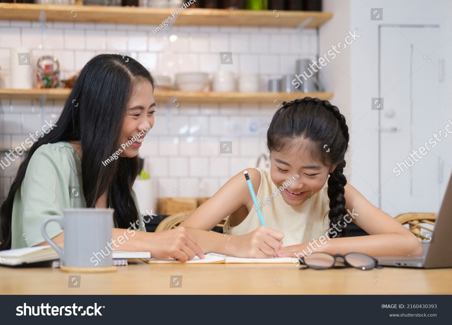 Smiling asian mom helping her sweet little daughter doing homework in home kitchen.
