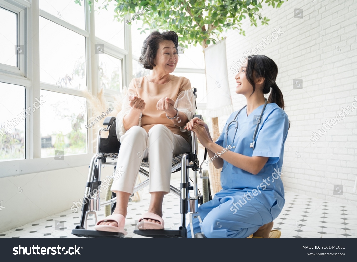 Young Asian nurse talking to a smiling senior female patient on a wheelchair and comforting her.